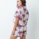 Woman wearing a shirt with pink palm trees and a matching bottom. She's standing behind a white background. The purpose of this image is to highlight the clothing by Mar de Lua.