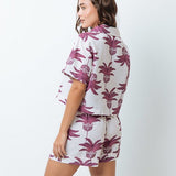 Woman wearing a shirt with pink palm trees and a matching bottom. She's standing behind a white background. The purpose of this image is to highlight the clothing by Mar de Lua.