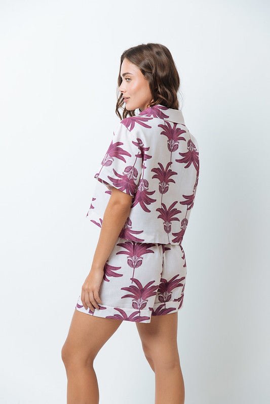 Woman wearing a shirt with pink palm trees and a matching bottom. She's standing behind a white background. The purpose of this image is to highlight the clothing by Mar de Lua.