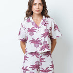 Woman wearing a shirt with pink palm trees and a matching bottom. She's standing behind a white background. The purpose of this image is to highlight the clothing by Mar de Lua.
