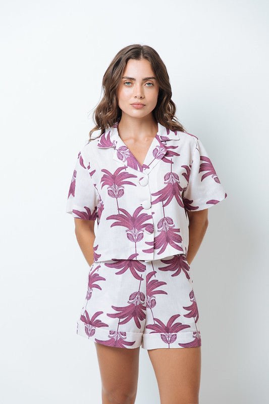 Woman wearing a shirt with pink palm trees and a matching bottom. She's standing behind a white background. The purpose of this image is to highlight the clothing by Mar de Lua.