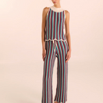 A woman modeling a sleeveless knit jumpsuit with vertical multicolor stripes in red, blue, green, and white, featuring a cream mock neck and drawstring waist, set against a solid light beige background.