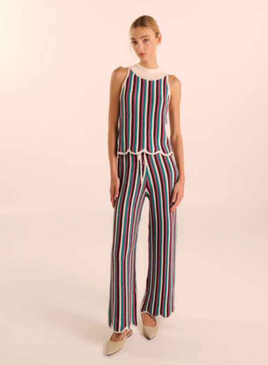 A woman modeling a sleeveless knit jumpsuit with vertical multicolor stripes in red, blue, green, and white, featuring a cream mock neck and drawstring waist, set against a solid light beige background.