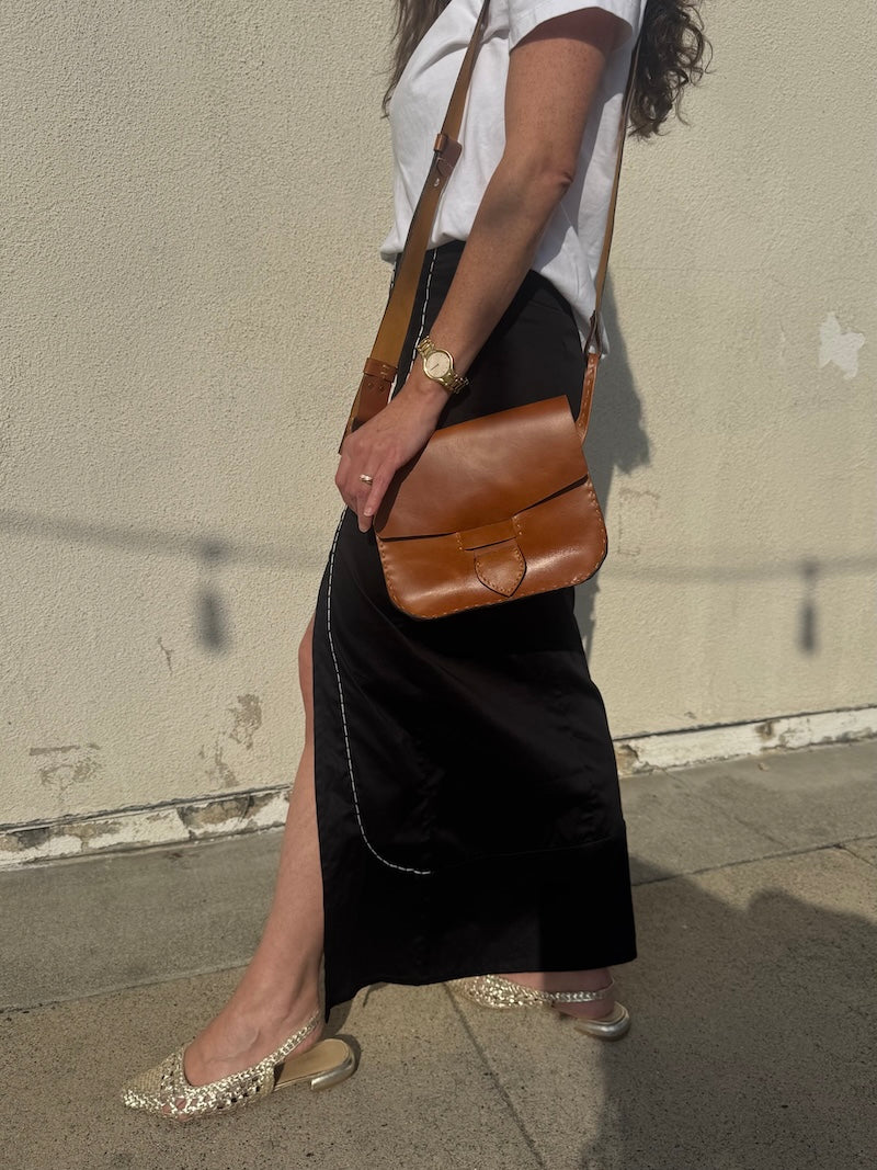 Person wearing a black skirt and white top with a brown leather bag against a plain wall.