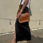 Person wearing a black skirt and white top with a brown leather bag against a plain wall.