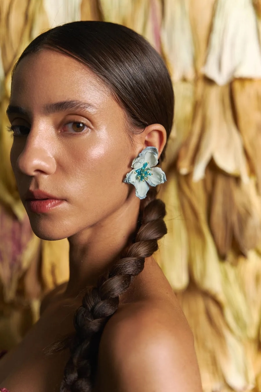 Woman with braided hair wearing floral earrings against a textured wall.