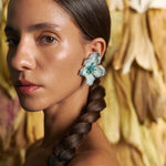 Woman with braided hair wearing floral earrings against a textured wall.