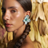 Woman with braided hair wearing floral earrings against a textured wall.