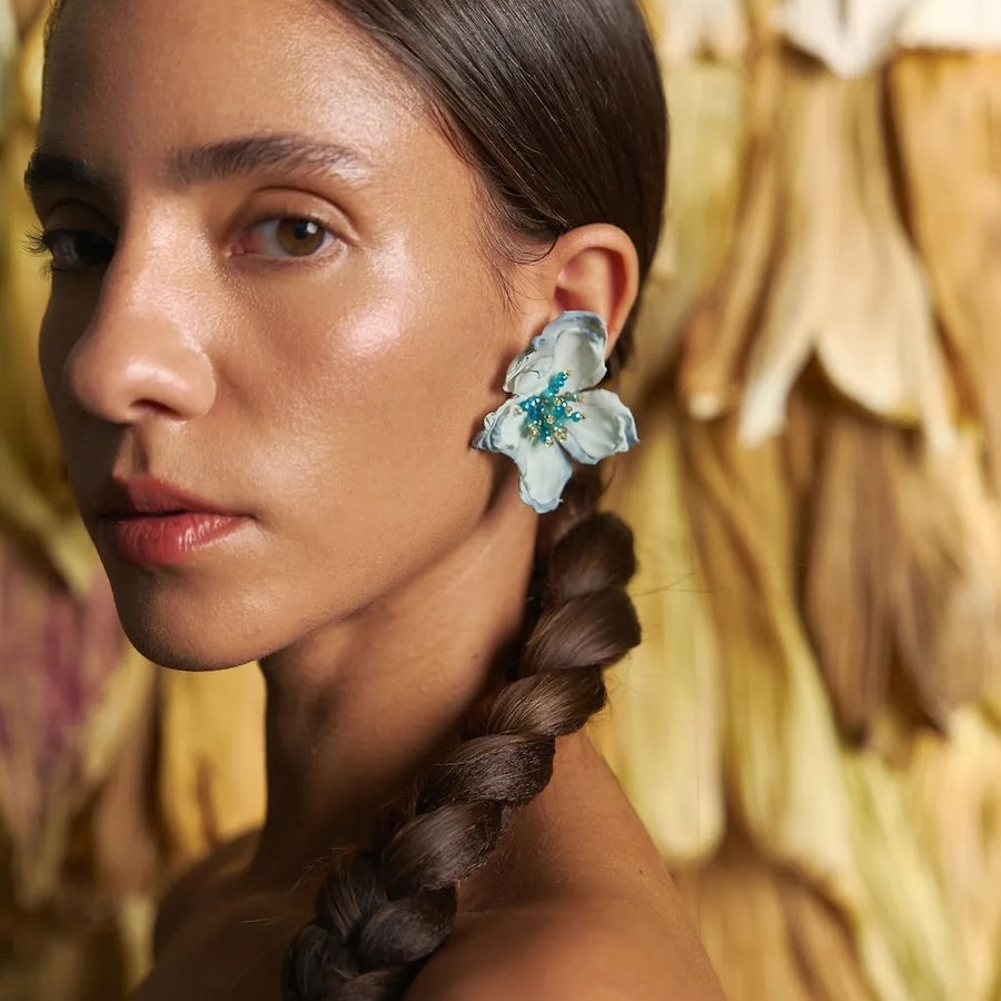 Woman with braided hair wearing floral earrings against a textured wall.