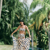 Woman wearing a white swimsuit with blue polka dots. She's holding a matching sarong and standing along the edge of a pool. The purpose of this image is to highlight the swimsuit and sarong because they're for sale at EVAMAIA Boutique.