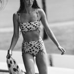 Black and white image of a woman wearing a swimsuit with polka dots. She's holding a matching sarong and walking along the edge of a pool. The purpose of this image is to highlight the swimsuit and sarong because they're for sale at EVAMAIA Boutique.