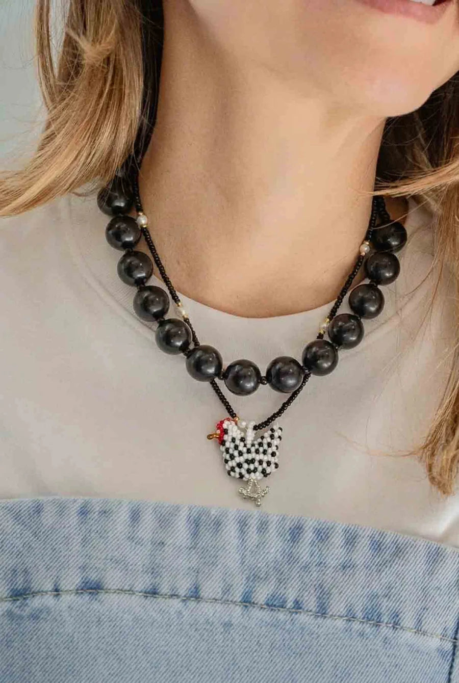 A close-up of a woman wearing a black beaded necklace with a black-and-white speckled rooster pendant and small red and white bead accents, paired with a white top and denim bottoms.
