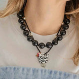 A close-up of a woman wearing a black beaded necklace with a black-and-white speckled rooster pendant and small red and white bead accents, paired with a white top and denim bottoms.