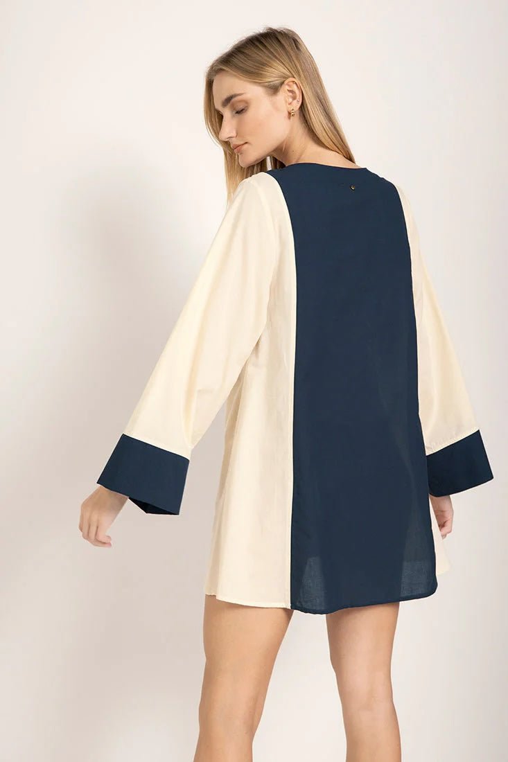 Image of the back of a model wearing a long sleeve color-block dress with white and navy tones. She's looking to the side and standing against a white background. The purpose of this image is to highlight the dress.
