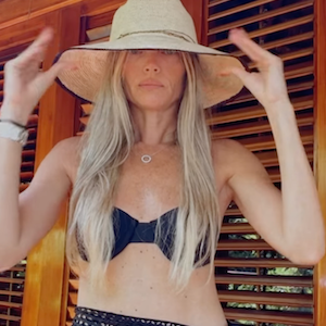 Woman wearing a wide-brimmed hat, black bikini top, and navy blue skirt against a wooden background.