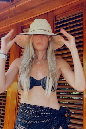 Woman wearing a wide-brimmed hat, black bikini top, and navy blue skirt against a wooden background.