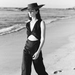 Black and white image of a woman wearing a black hat and a black jumpsuit with a cutout on the front. She's walking on the sand at the beach. The purpose of this image is to highlight the jumpsuit, which is on sale at EVAMAIA Boutique in Studio City, CA.