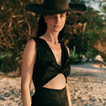 Image of a woman wearing a black hat and a black jumpsuit with a cutout on the front. She's standing on the sand at the beach. The purpose of this image is to highlight the jumpsuit, which is on sale at EVAMAIA Boutique in Studio City, CA.