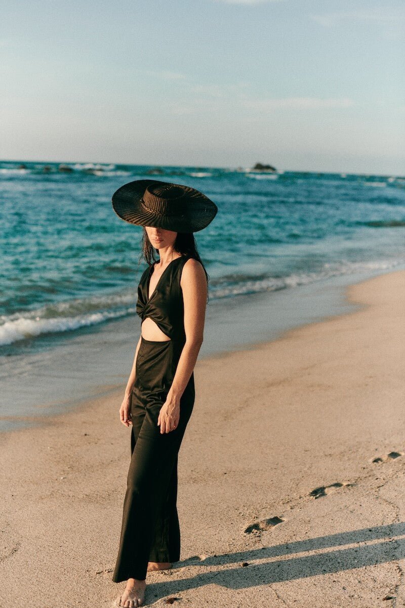 Image of a woman wearing a black hat and a black jumpsuit with a cutout on the front. She's standing on the sand at the beach. The purpose of this image is to highlight the jumpsuit, which is on sale at EVAMAIA Boutique in Studio City, CA.