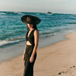 Image of a woman wearing a black hat and a black jumpsuit with a cutout on the front. She's standing on the sand at the beach. The purpose of this image is to highlight the jumpsuit, which is on sale at EVAMAIA Boutique in Studio City, CA.