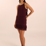 A woman modeling a sleeveless, fringed mini dress in a deep burgundy or maroon shade, paired with olive green flat sandals, against a solid light beige studio background.