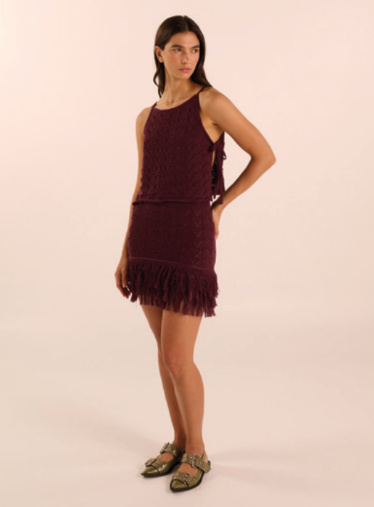 A woman modeling a sleeveless, fringed mini dress in a deep burgundy or maroon shade, paired with olive green flat sandals, against a solid light beige studio background.