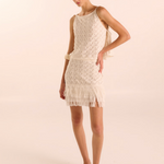 A woman modeling a sleeveless cream crochet mini dress with fringe trim and a matching shoulder bag, paired with beige pointed-toe flats, against a solid light beige studio background.