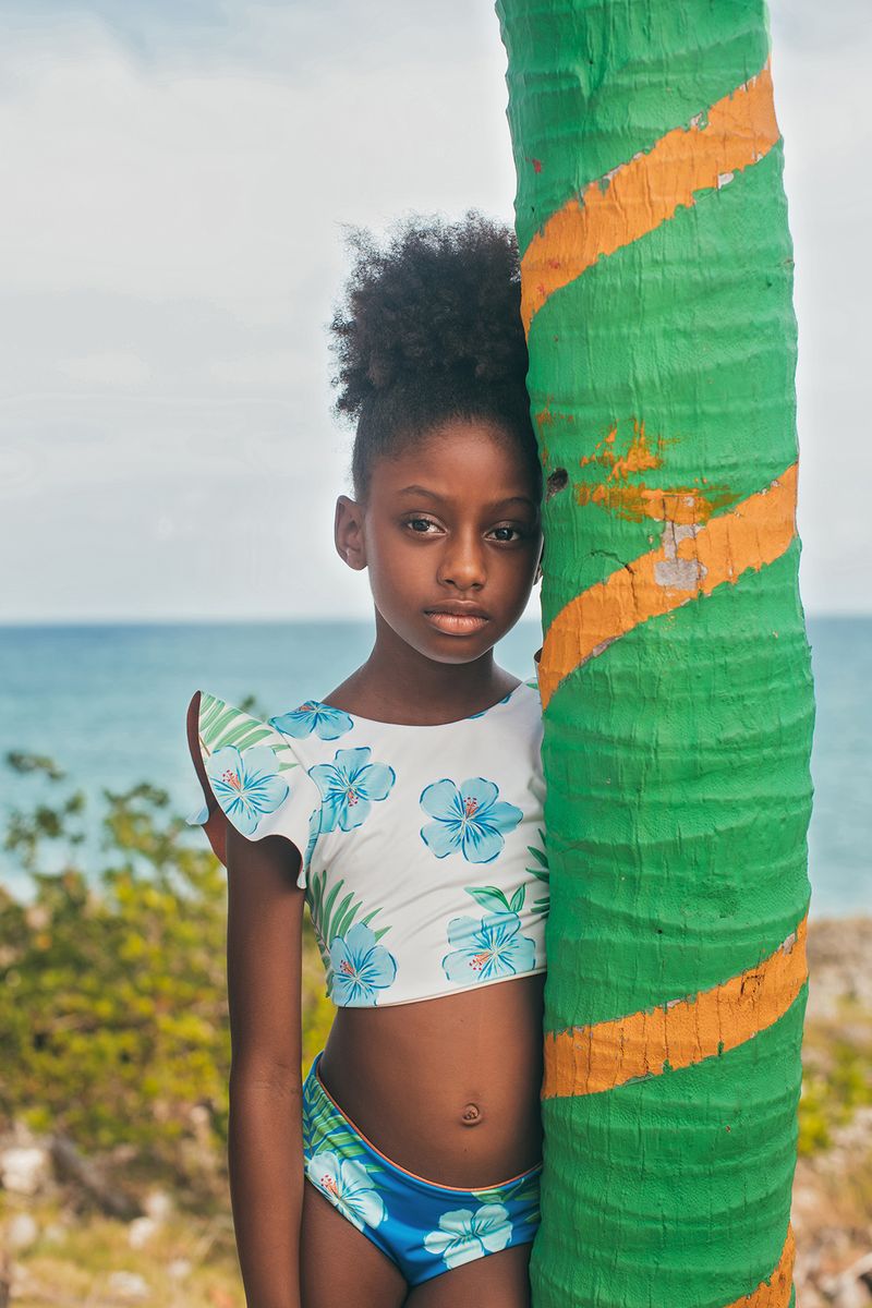 Young girl in a floral swimsuit standing next to a colorful palm tree sculpture with ocean in the background