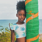 Young girl in a floral swimsuit standing next to a colorful palm tree sculpture with ocean in the background