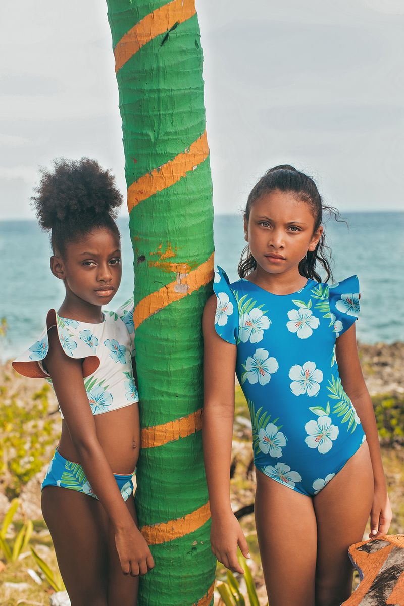 Two young girls in floral swimsuits standing next to a palm tree with ocean in the background