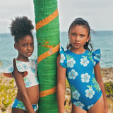 Two young girls in floral swimsuits standing next to a palm tree with ocean in the background