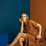 Image of a woman wearing sunglasses on top of her head and an embroidered set. She is sitting against a brown and blue background. She's looking at the camera. The purpose of this image is to highlight the skirt and shirt.
