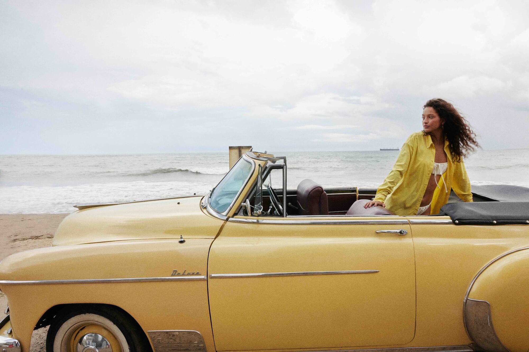 A woman in a yellow shirt leans out of a vintage yellow convertible car parked on a beach with the ocean and cloudy sky in the background.