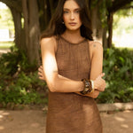 Woman in a brown crochet dress standing outdoors with greenery in the background.
