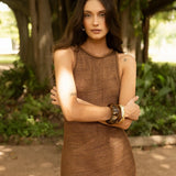 Woman in a brown crochet dress standing outdoors with greenery in the background.