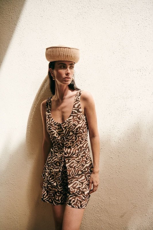 Woman wearing brown shorts and a matching vest. She's looking to the side, in a pensive way. She's also wearing a small straw hat.