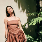 Image of a model wearing a brown dress with Spaghettin straps. She's standing against a wall and seems tired.