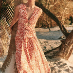 Woman wearing an orange and tan dress. She's standing near a palm tree on the beach. The purpose of this image is to highlight the dress by Encantadore Swim and Resortwear.