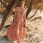 Woman wearing an orange and tan dress. She's standing near a palm tree on the beach. The purpose of this image is to highlight the dress by Encantadore Swim and Resortwear.