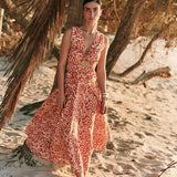 Woman wearing an orange and tan dress. She's standing near a palm tree on the beach. The purpose of this image is to highlight the dress by Encantadore Swim and Resortwear.