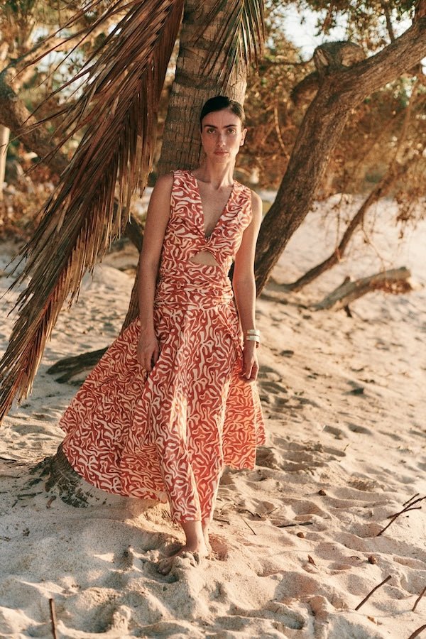 Woman wearing an orange and tan dress. She's standing near a palm tree on the beach. The purpose of this image is to highlight the dress by Encantadore Swim and Resortwear.