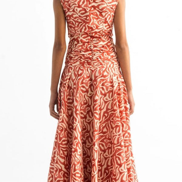 Back of a woman wearing an orange and tan dress. She's standing on a white background. The purpose of this image is to highlight the dress by Encantadore Swim and Resortwear.