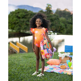 A sweet little girl posing with this cute orage sun one piece swimsuit. There a small picnic set up behind her!