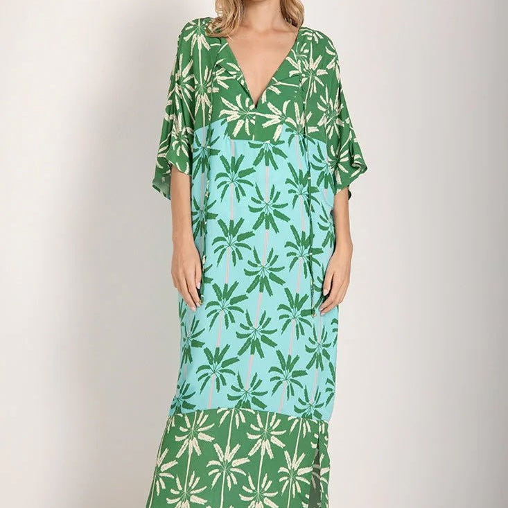 Woman wearing a long cover-up dress with a green and blue palm tree print. She's looking towards the camera and standing in front of a white background. The purpose of this image is to highlight the dress.