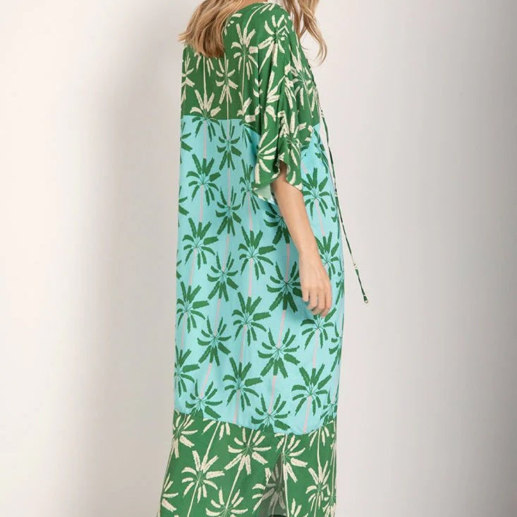 Woman wearing a long cover-up dress with a green and blue palm tree print. She's looking down and to the side. Her body is turned away from the camera.  She's standing against a white background. The purpose of this image is to highlight the dress.