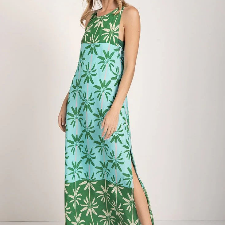 Woman wearing a sleeveless long cover-up dress with a green and blue palm tree print. She's looking towards the camera and standing in front of a white background. The purpose of this image is to highlight the dress.