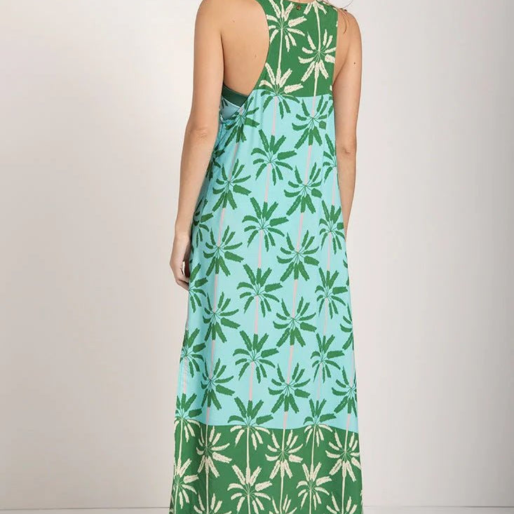 Woman wearing a sleeveless long cover-up dress with a green and blue palm tree print. She's looking to the side and standing against a white background. The purpose of this image is to highlight the dress.