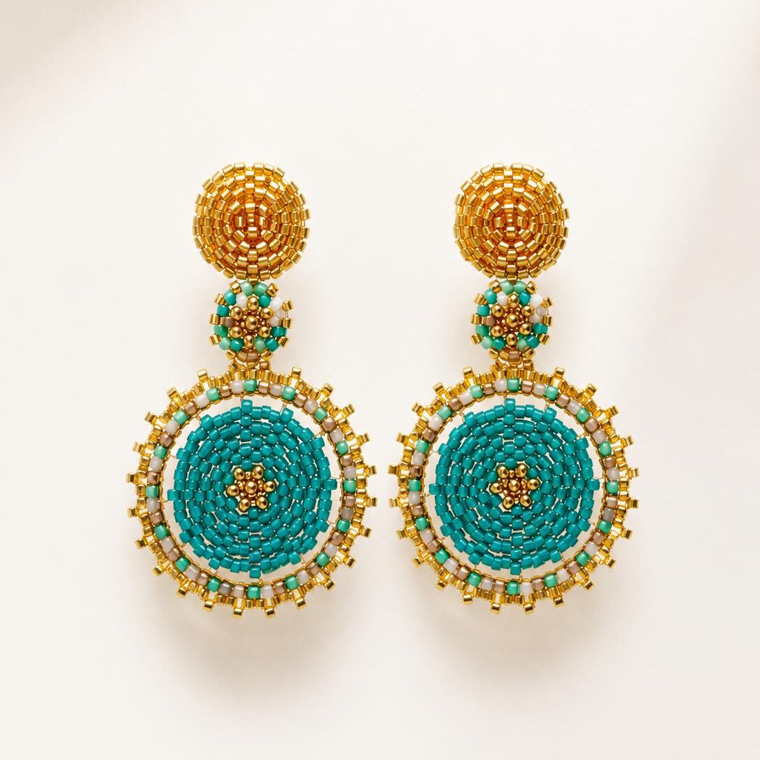 A pair of ornate handmade beaded drop earrings with gold-toned metal posts and circular turquoise, white, and gold beadwork, displayed on a clean off-white background.