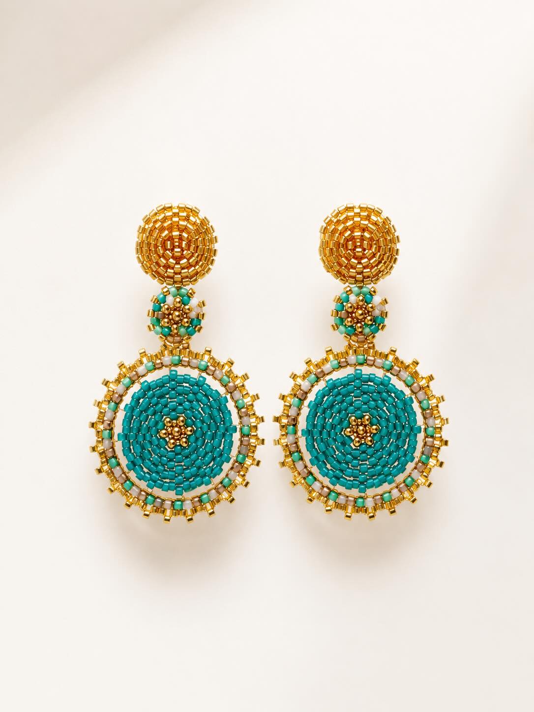 A pair of ornate handmade beaded drop earrings with gold-toned metal posts and circular turquoise, white, and gold beadwork, displayed on a clean off-white background.