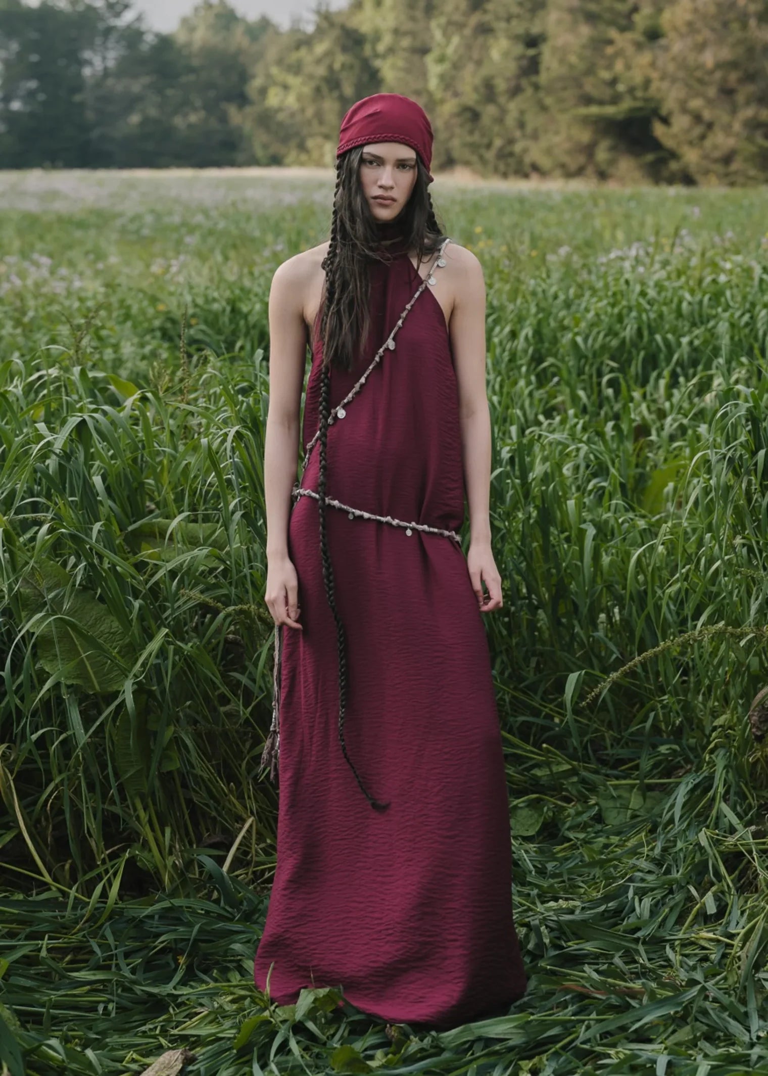 Woman in a long burgundy dress and red headscarf standing in a field of tall grass.